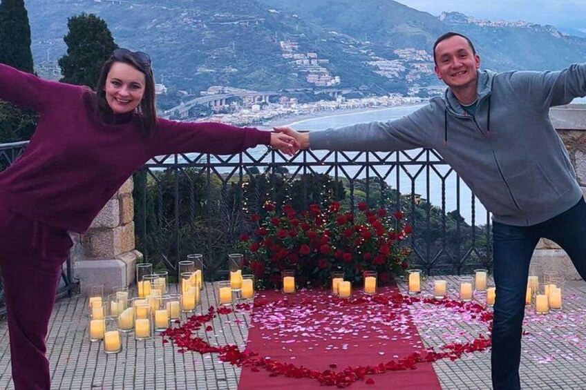 Two people holding hands on a balcony overlooking a scenic landscape with a table decorated with candles and rose petals. 2