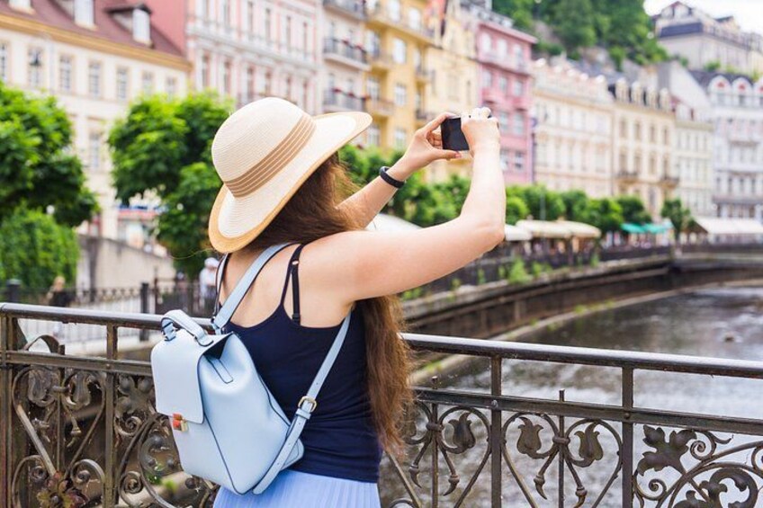Une personne prend une photo avec un téléphone intelligent, porte un chapeau et une robe bleue, et se tient près d’une rivière, avec des bâtiments en arrière-plan. 5