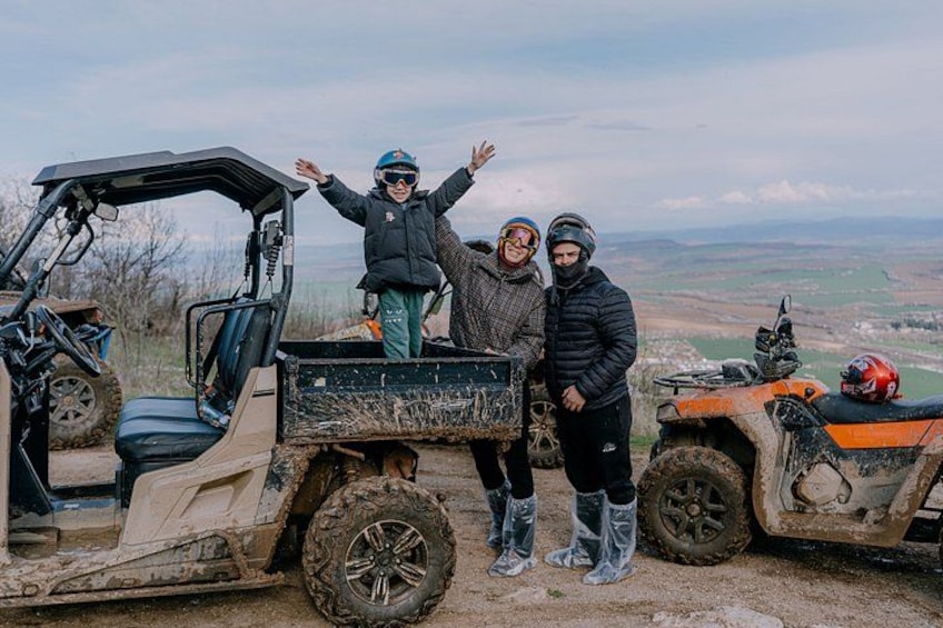 Three people standing on a dirt path with off-road vehicles, a clear sky, and a hilly landscape in the background. 1