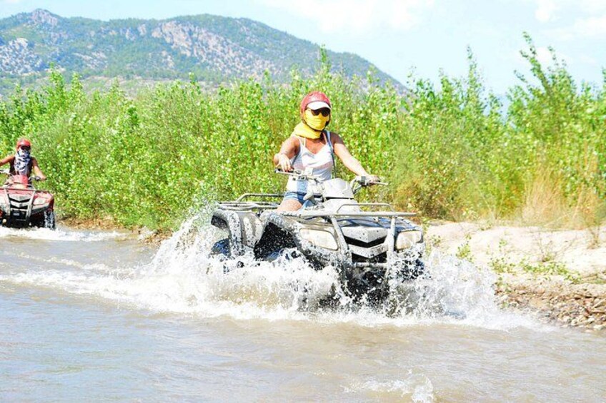 Two people riding ATVs on a river surrounded by greenery and mountains. 3