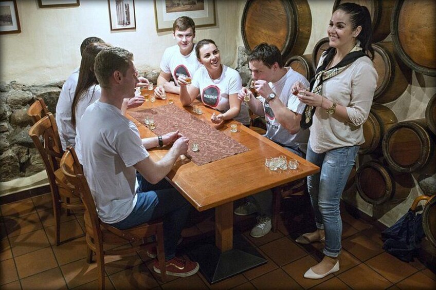 Un gruppo di persone attorno a un tavolo di legno con bicchieri, in una stanza piena di botti di vino. 4