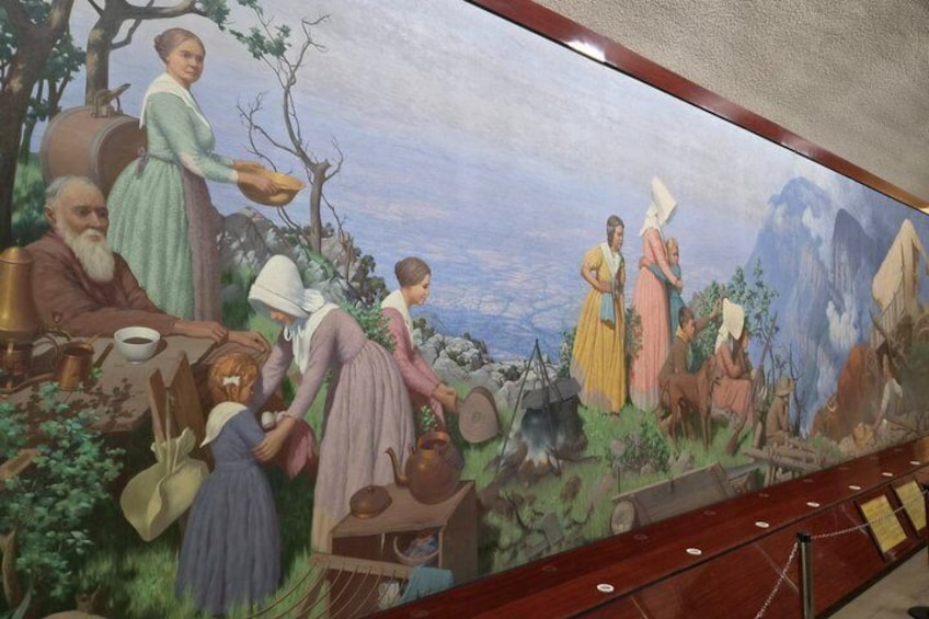 Un mural que representa una escena histórica con personas vestidas con ropa de época, una mesa con comida y un paisaje montañoso. 3