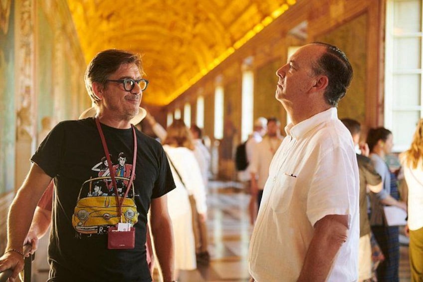 A person in a black t-shirt with a graphic design and a lanyard, standing in a corridor with a high ceiling and columns. 3