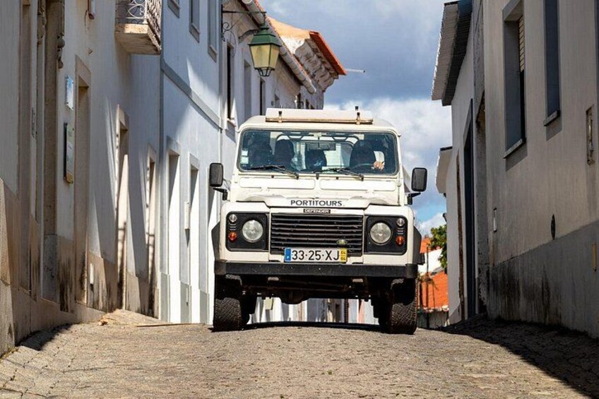 Un Land Rover blanc roule sur une rue pavée, entre deux bâtiments. 2