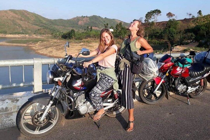 Dos personas sentadas en motocicletas a orillas de un lago, con colinas al fondo. 1
