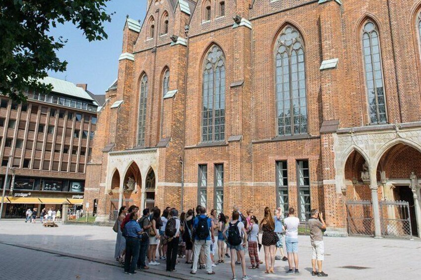 A group of people standing in front of a large brick building with arched windows and a clock tower. 5