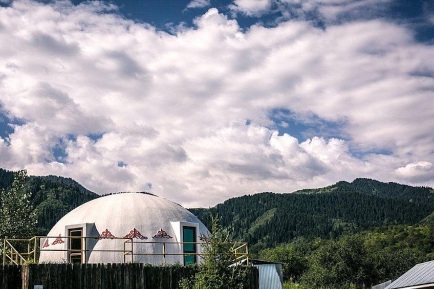 A white dome-shaped structure with a green door, surrounded by a wooden fence, set against a backdrop of a forested mountain range under a partly cloudy sky. 4
