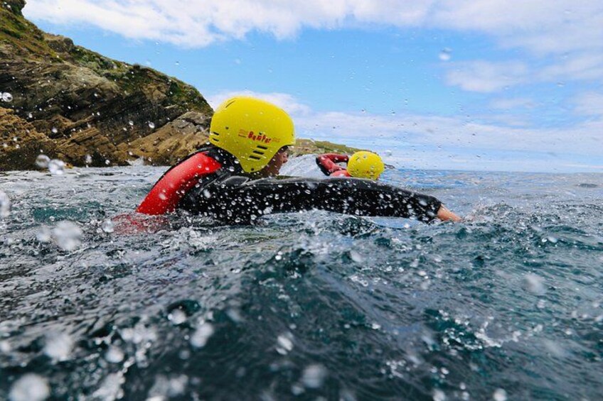 Two people in a kayak wearing helmets, paddling in a body of water with a rocky shoreline in the background. 1