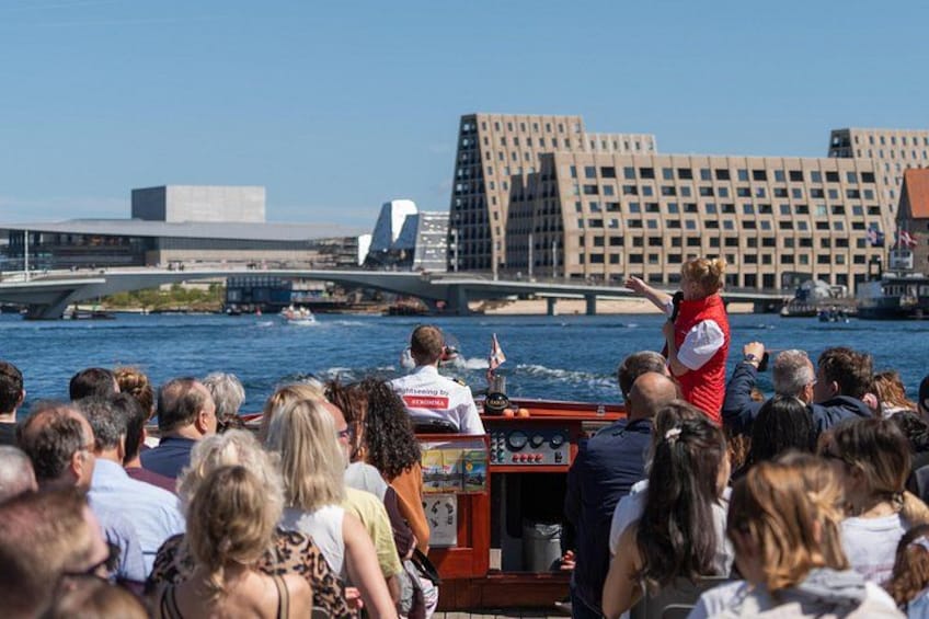 Une foule à bord d’un bateau, une personne qui prend la parole devant l’assistance, et un bâtiment moderne en arrière-plan. 2