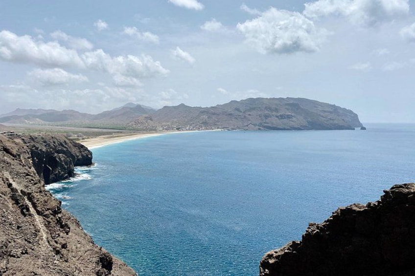 A coastal landscape with a clear blue sea, rocky cliffs, and distant mountains under a partly cloudy sky. 2