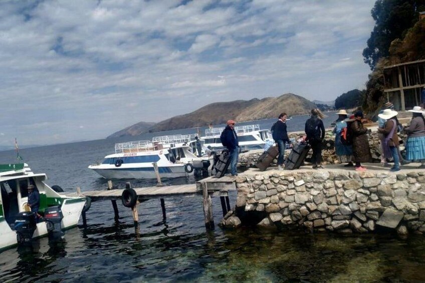 Eine Steg mit vor Anker liegenden Booten, Personen auf dem Steg und eine gebirgige Landschaft im Hintergrund. 1