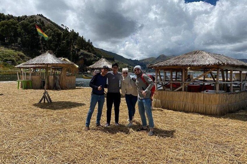 A group of people standing on a straw-covered ground with thatched-roof structures and a flag in the background. 3