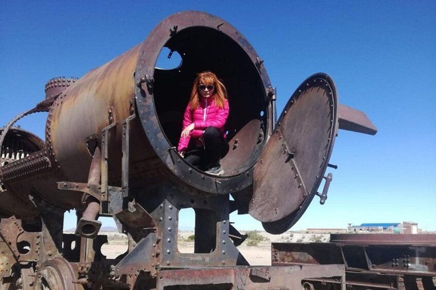 A person in a pink jacket sitting inside a large, rusted cylindrical structure with open ends, outdoors. 3