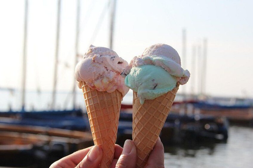 Due coni gelato tenuti da una persona, con uno sfondo che include un porto turistico e barche. 4