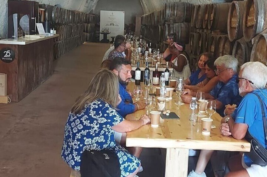 Un grupo de personas sentadas a una larga mesa de madera en una bodega, rodeadas de barriles de vino. 1