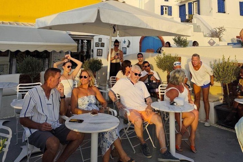 People sitting at outdoor tables, white umbrellas, and a building with blue shutters in the background. 4