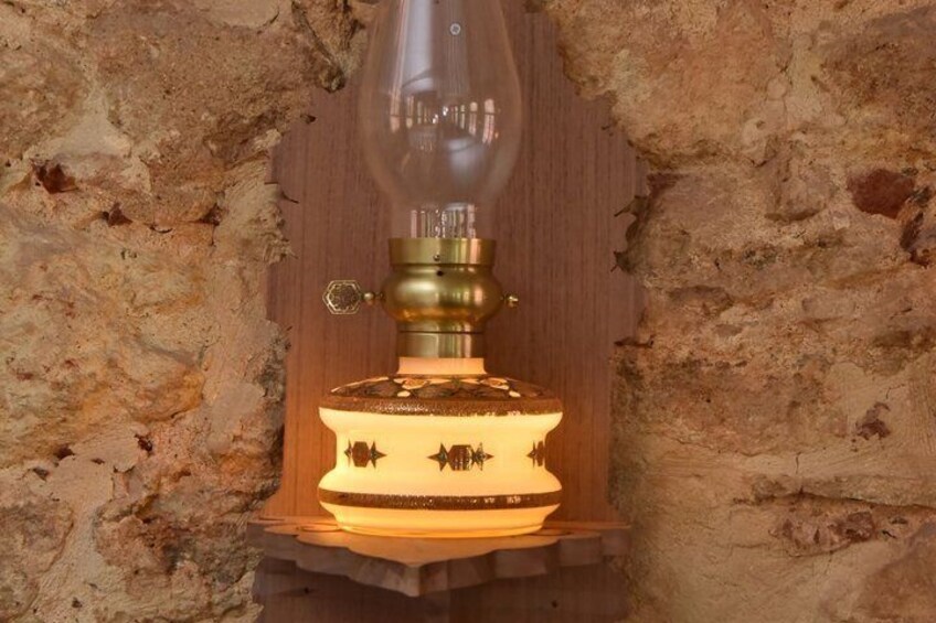 A vintage oil lamp with a brass base and a glass chimney, mounted on a wooden stand against a textured stone wall. 4