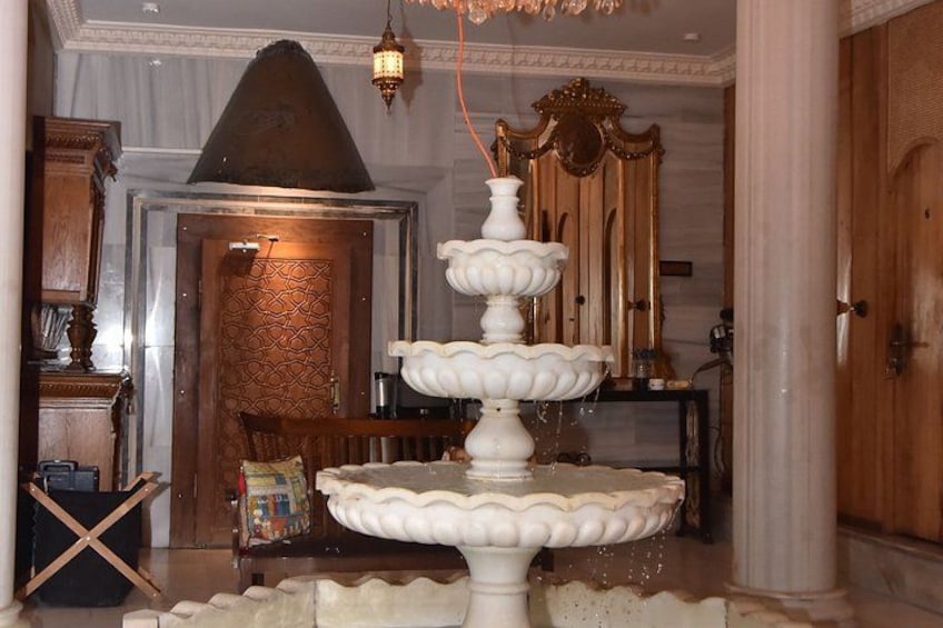 A multi-tiered white fountain in a room with a wooden door, a chandelier, and a mirror. 3