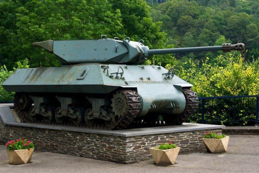 A military tank on display, surrounded by greenery and stone structures. 1