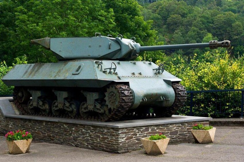 A military tank on display, surrounded by greenery and stone structures. 1