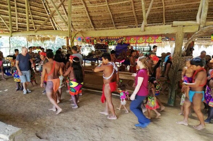A group of people in a covered outdoor area with a thatched roof, some wearing colorful traditional attire. 3