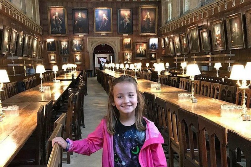 A long dining hall with wooden tables and chairs, and framed portraits on the walls. 3