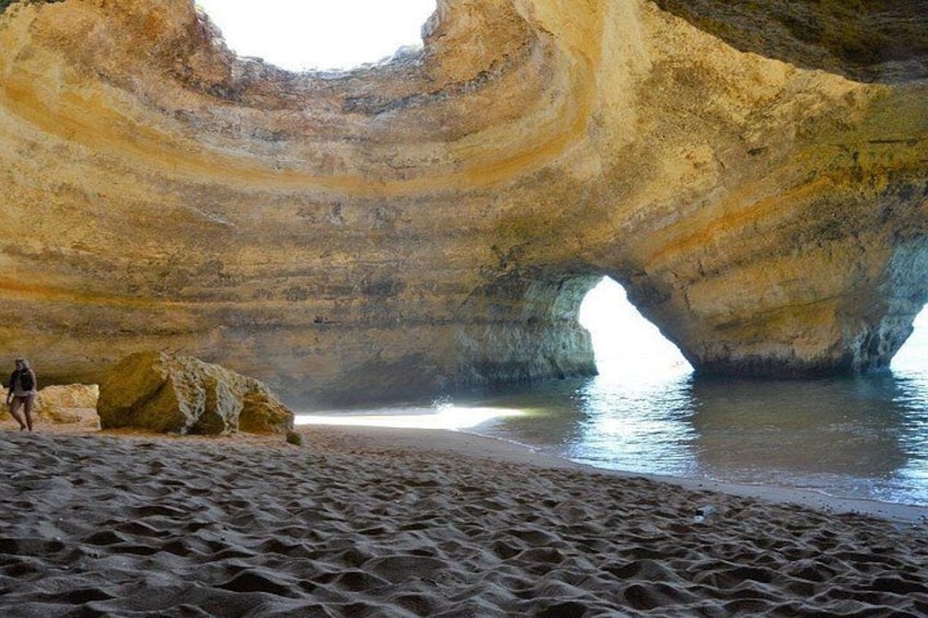 Eine Person geht an einem sandigen Strand entlang, mit einer großen Felsformation und einem hohlen Durchgang. 2