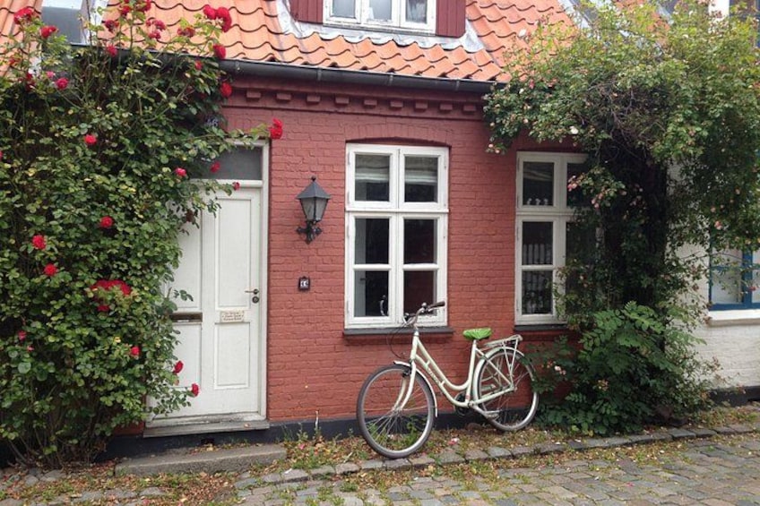 Una casa de ladrillo rojo con una puerta blanca, una bicicleta estacionada adelante y un rosal con flores rojas. 3