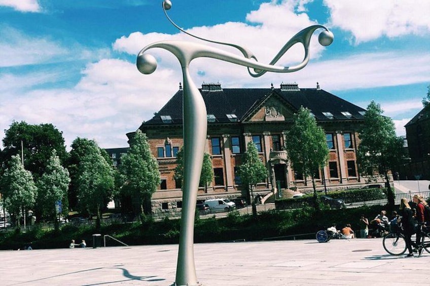 A public square with a modern sculpture, a building with a brick facade, and people walking and cycling. 2