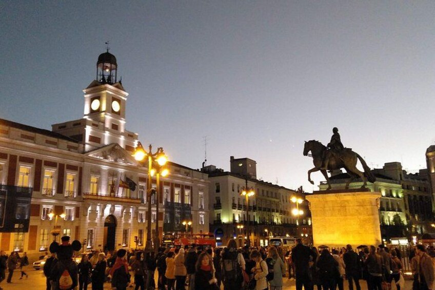 A public square with a statue of a mounted figure, a clock tower, and illuminated buildings. 5