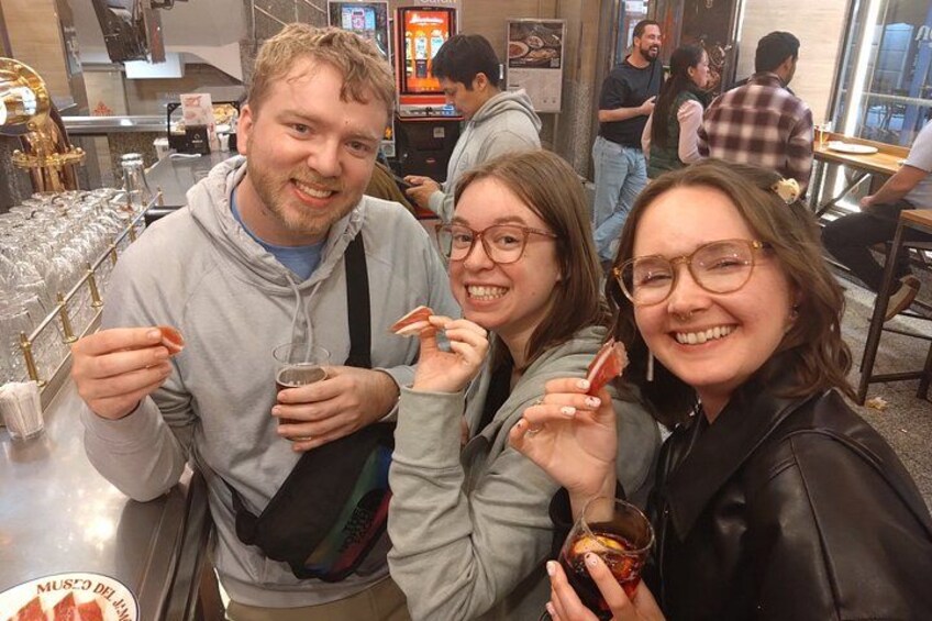 Three people holding food items and drinks in a casual dining setting with a counter and other diners in the background. 4