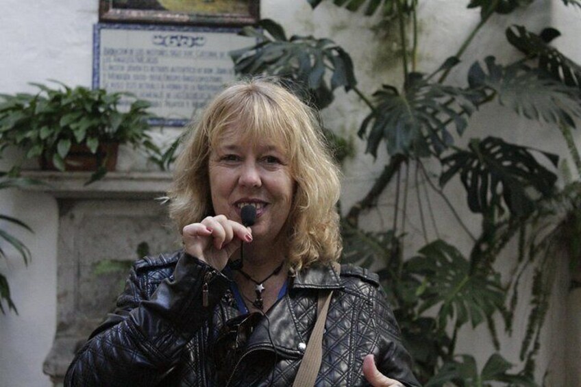 Una persona con chaqueta de cuero y un micrófono, parada frente a una pared con una placa y plantas tropicales. 5