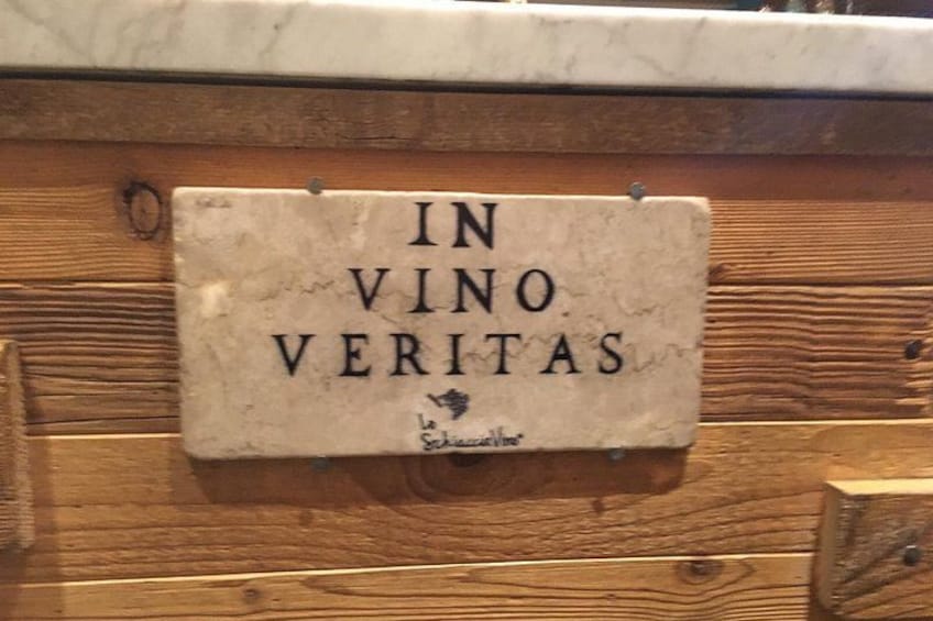 Een houten wand met een bord waarop staat 'IN VINO VERITAS' en een klein handgeschreven briefje eronder. 4