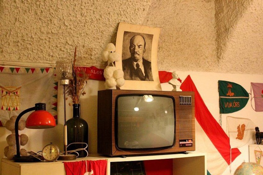 A vintage television on a shelf, a red lamp, a vase with dried plants, a framed picture, and a wall with flags and decorations. 5