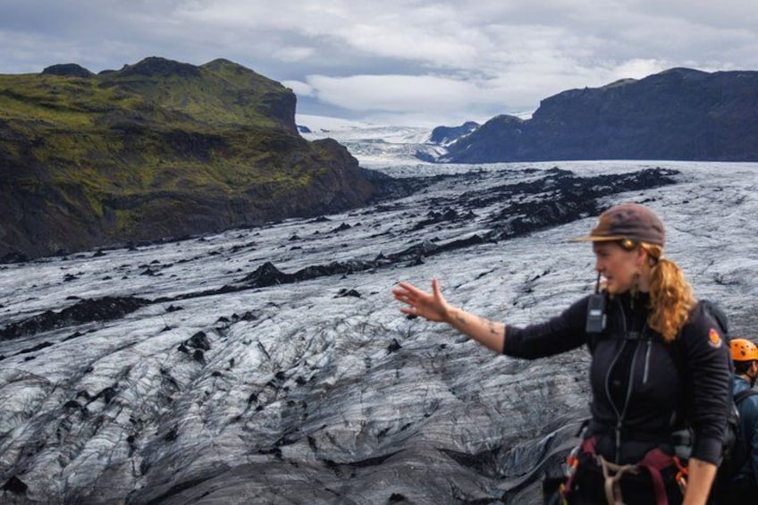 A person in a black jacket and helmet, gesturing towards a glacier with a backdrop of mountains. 3