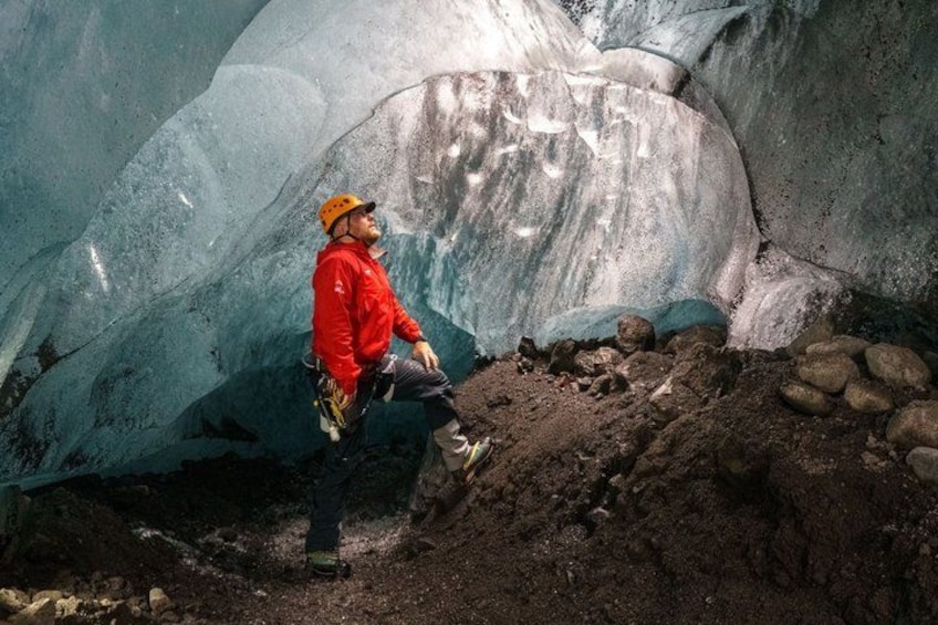 A person in a red jacket and yellow helmet sitting on a rocky ledge with a glacier in the background. 3