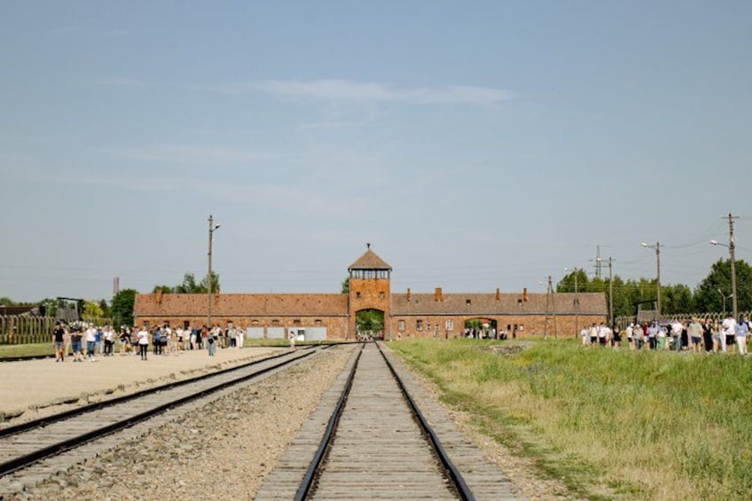 A railway track leading to a brick building with a tower, surrounded by a crowd of people. 1