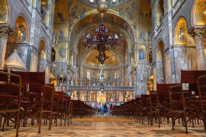 Een versierd kerkinterieur met hoge plafonds, gedetailleerde fresco's en een centrale kroonluchter. Rijen houten banken flankeren de gangpaden. 2