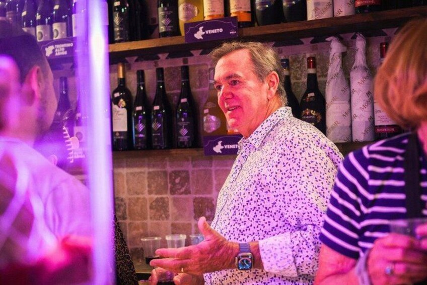 Une personne portant une chemise à motifs, debout dans un bar, avec des étagères remplies de bouteilles en arrière-plan. 4