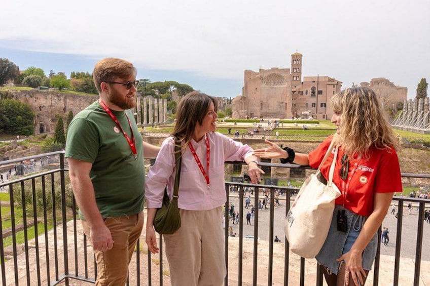 Three people standing by a railing, overlooking ruins and a large building in the background. 2