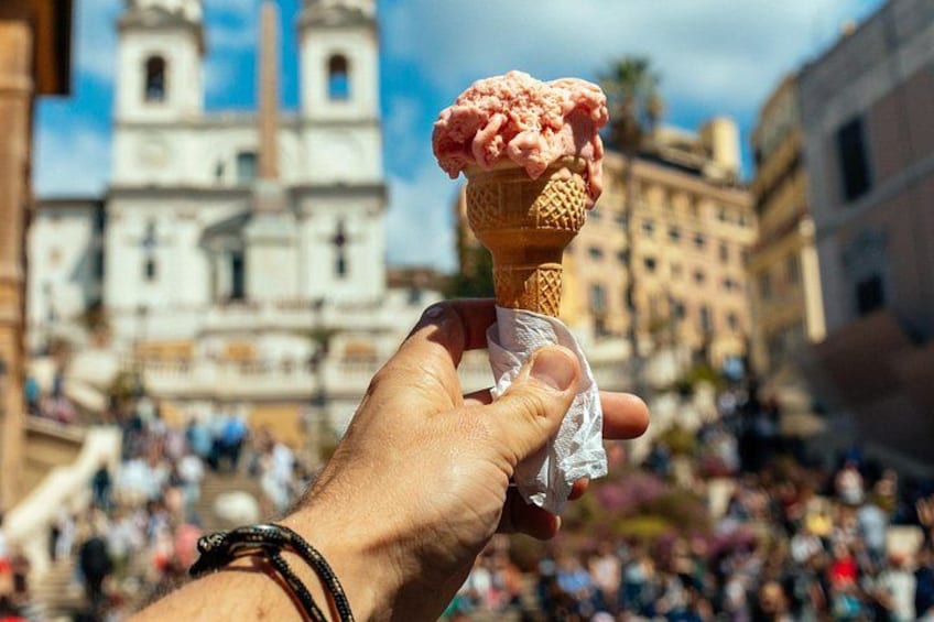 A hand holding an ice cream cone with a scoop of pink ice cream in front of a historic building with a bell tower. 1