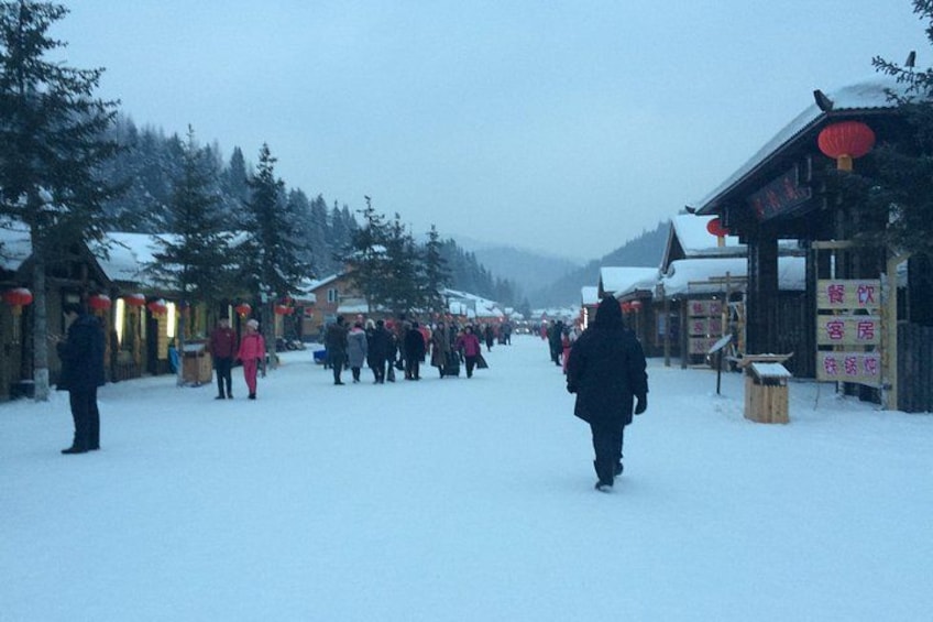 A snow-covered street with people walking, buildings with traditional architecture, and trees lining the street. 3