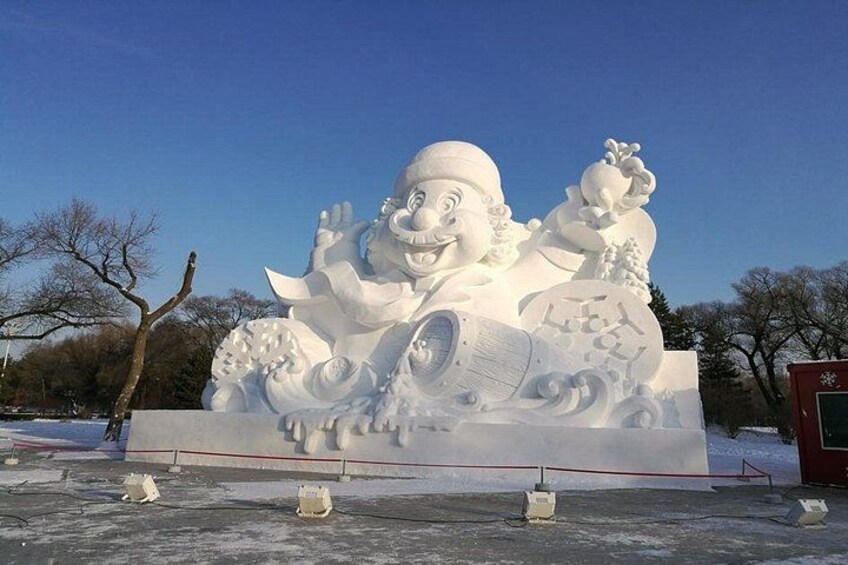 A large snow sculpture of a smiling figure with a hat and a cane, surrounded by smaller figures and text. 2