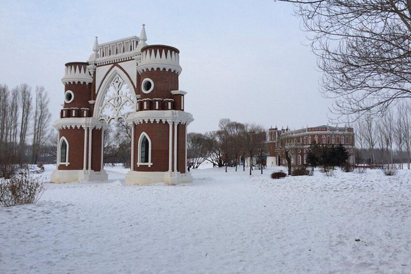 A snow-covered architectural structure with a gothic arch and brickwork, surrounded by bare trees and a distant building. 3