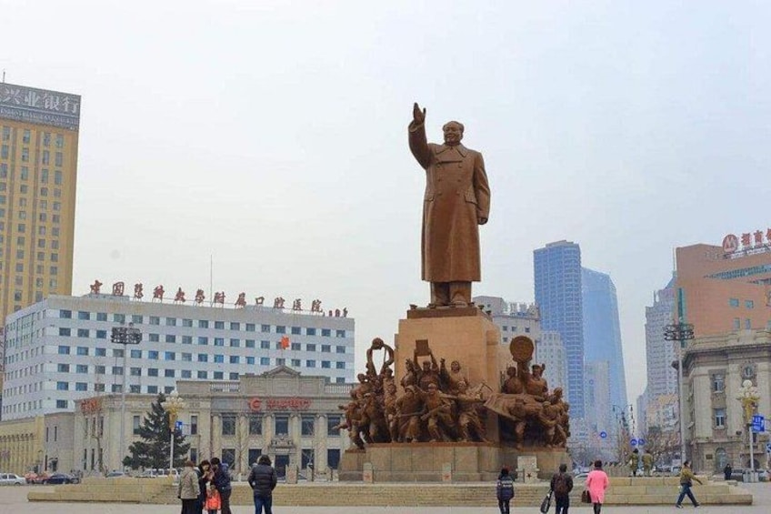 A large statue of a man in a coat, surrounded by a crowd of people, with modern buildings in the background. 1