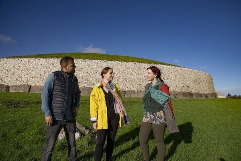 Three people standing in front of a large stone structure with a grassy field and a clear sky in the background. 3