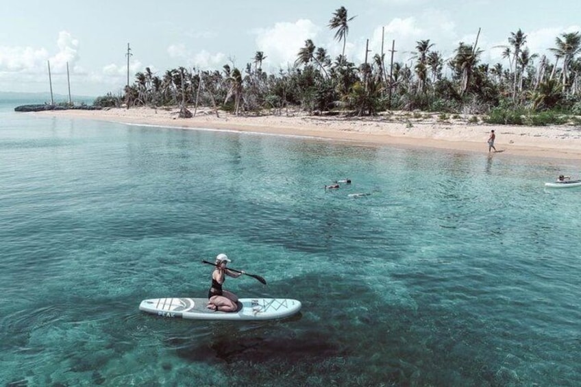 A person paddleboarding on clear turquoise water with a beach and palm trees in the background. 2
