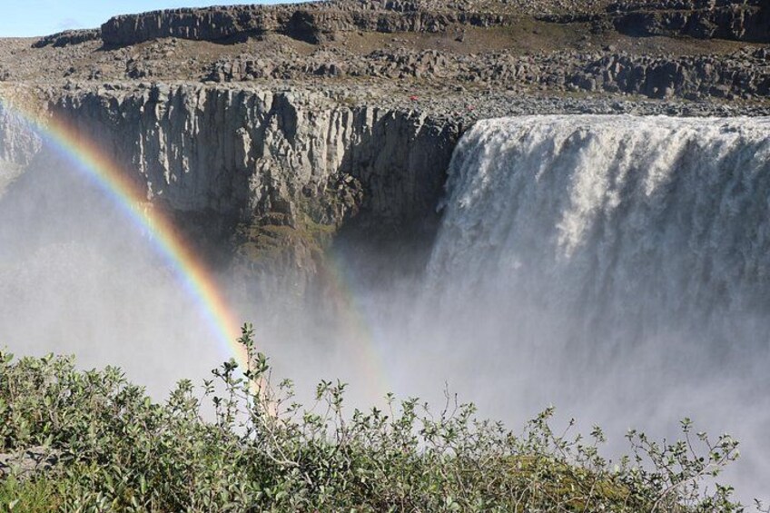 A waterfall with a rainbow and sparse vegetation in the foreground. 2