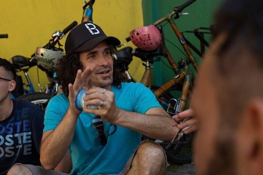 A person wearing a black cap with a white logo, sitting outdoors with bicycles and helmets in the background. 2