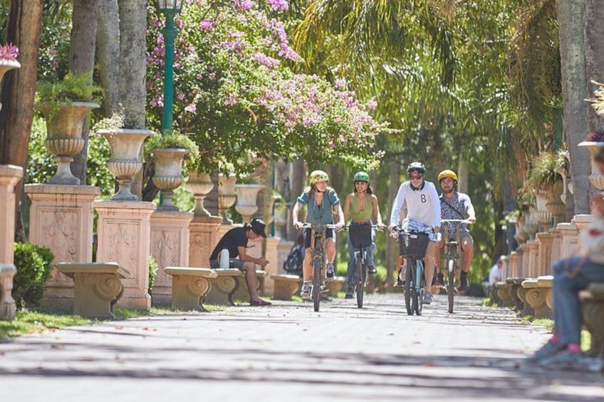 En grupp cyklister på en asfalterad stig, omgiven av klassisk arkitektur och blommande växter. 5
